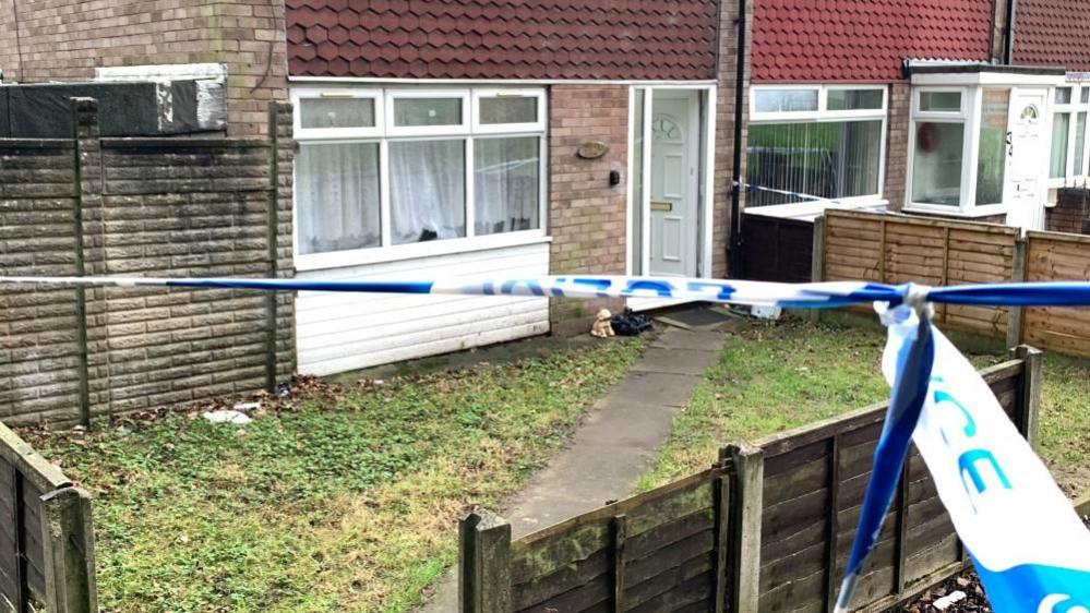 A blue police cordon blocks the entry to a pale-bricked house. The tape stretches across the image which shows a property with a narrow concrete path leading to a white plastic door that is partially open. There is grass on either side of the path and a low fence at the front of the house.