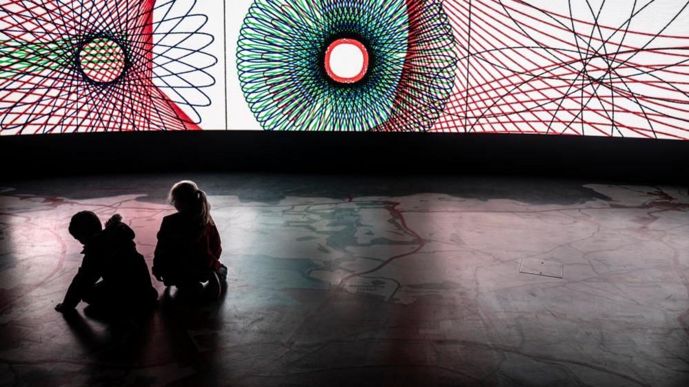 Tow childrensit on the floor of a darkened room while mathematical circular sprigrpah images are projected onto a lit wall.