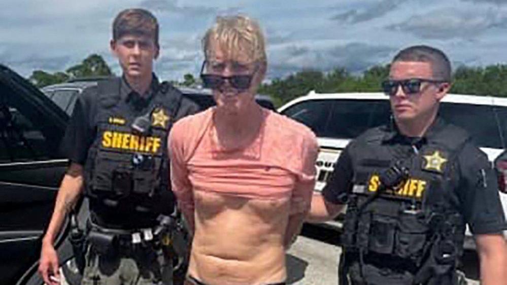 A still from a police video showing Ryan Routh, a man with a mop of blond hair, sunglasses and a pink T-shirt pulled up over his stomach, flanked by two deputies from the Martin County Sheriff's Office