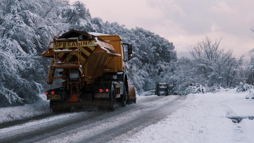 Two gritting trucks treat a snowy road surrounded by evergreen trees. The sky is cloudy.