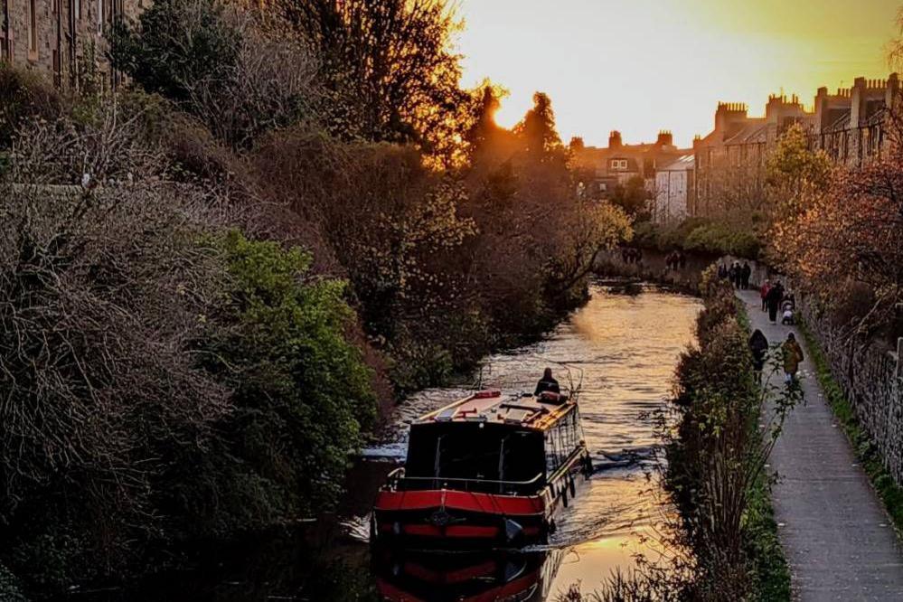 A narrowboat glides along a tree-lined canal as the sun sets behind rooftops.
