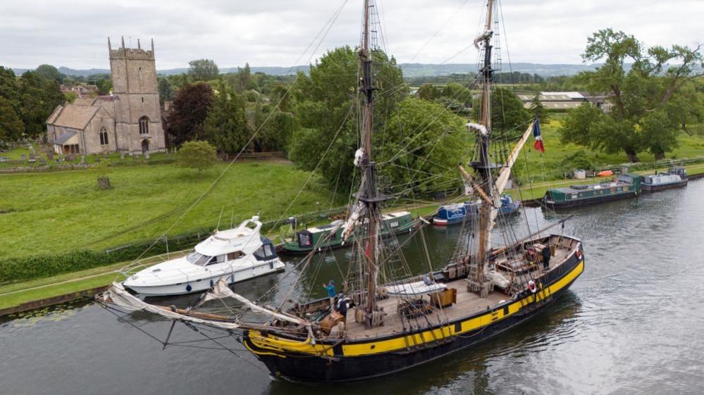 A tall-rigged ship sailing up the canal into Gloucester Docks. A church can be seen on the river bank behind it, and other smaller, more modern boats are moored on the river too