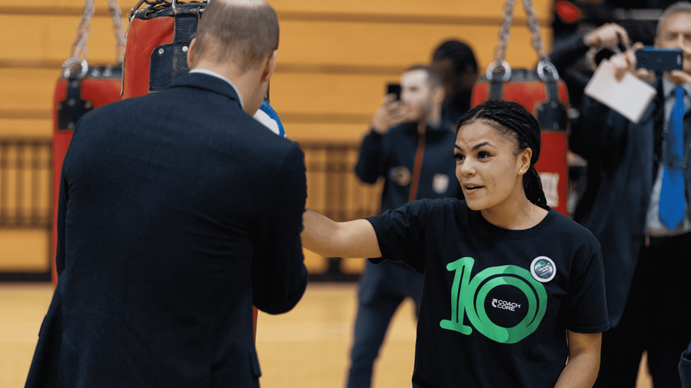 A girl, wearing a black and green Coach Core t-shirt, thrusts her left hand towards a man in a suit in a punching action.