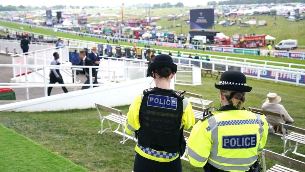 Two female police officers face toward Epsom Downs race course wearing high-vis uniforms labelled 'Police'.