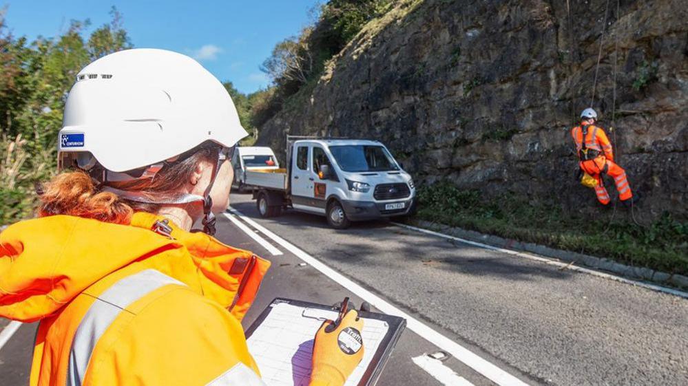 Council highways team check the rock face on the A170 on Sutton Bank. One man is suspended in a harness on the rock face while a female worker stands with a clipboard on the other side of the road