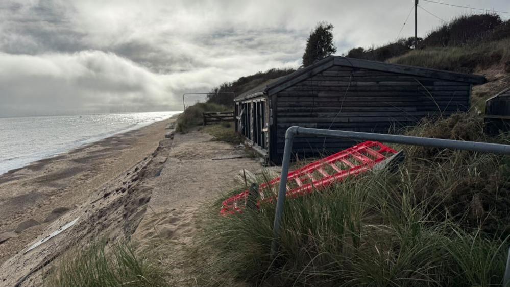 A black-painted timber clad chalet, with glazed frontage. The windows have been smashed. It sits aloft the cliff top, which slopes towards the left down to the beach. A metal hand rail and orange plastic fence panel are among the marram grass in the foreground.