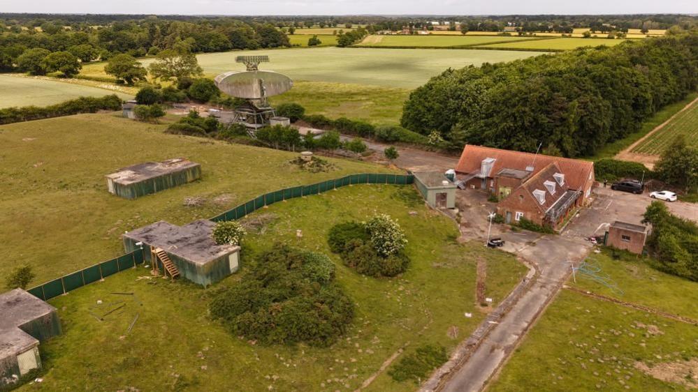 A drone image of part of the RAF Neatishead site, with bunker and ventilation access points and an office accommodation building. A concrete roadway runs through the site and green fencing can be seen sectioning off some areas. THere are green fields and trees, creating a woodland environment. In the distance is the T84 Radar, to the upper left of the image.