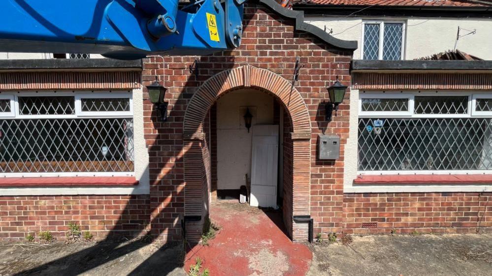The entrance of the Fritton Decoy Tavern was built in the 1930s to 1950s featuring a tiled column and arch porch, with a Dutch gable over the top. Bay windows feature either side, with magnolia render above. A blue cherry picker arm is visible in the upper left of the image.
