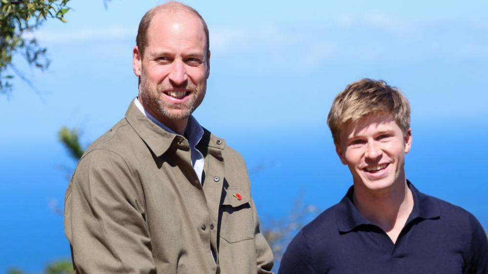 Prince William and Robert Irwin smile for a photo, a deep blue sky extending behind them