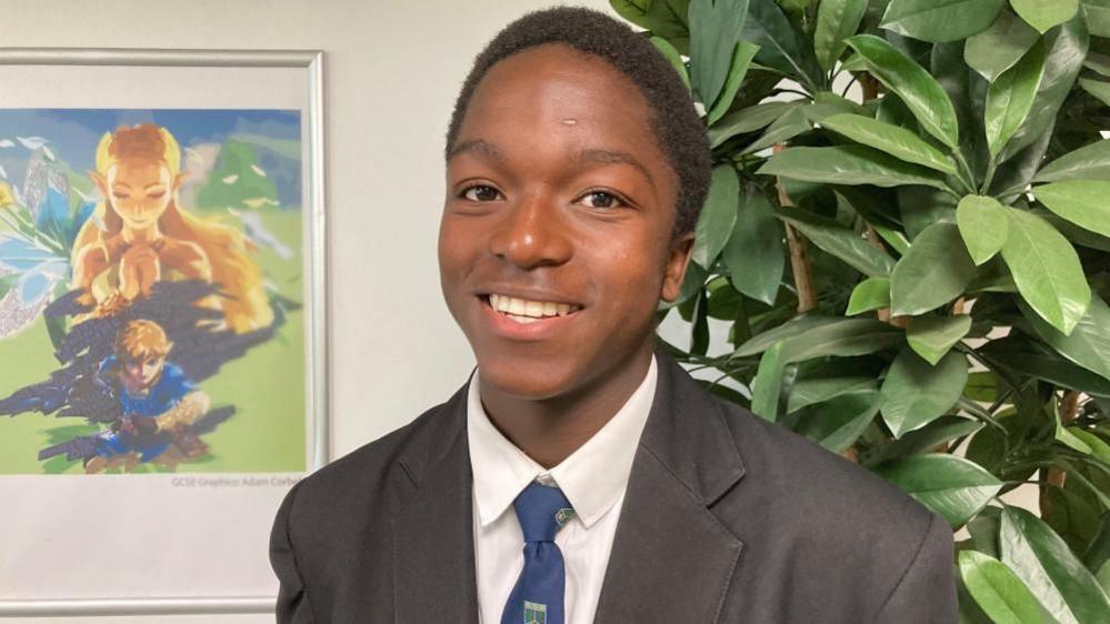 Nokutenda smiles at the camera wearing a school uniform, standing by a picture and a plant