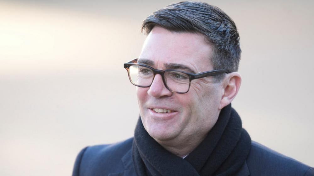 Greater Manchester Mayor Andy Burnham stands wearing a jacket and a black scarf and smiling. 