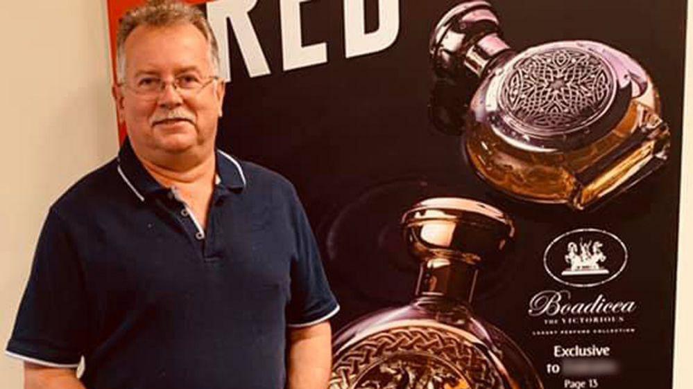 David Crisp, a man with short greying hair, a grey moustache and glasses, wearing a navy polo t-shirt and his hands clasped, stands in front of a poster showing two circular perfume bottles embossed with horses and a circular pattern and the Boadicea logo in italics. 