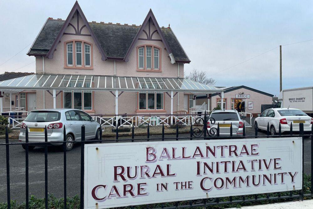 A large detached light pink house with a dark grey roof. Cars are parked in front of it and a black iron fence is in the foreground with a sign which says "Ballantrae Rural Initiative Care in the Community". 