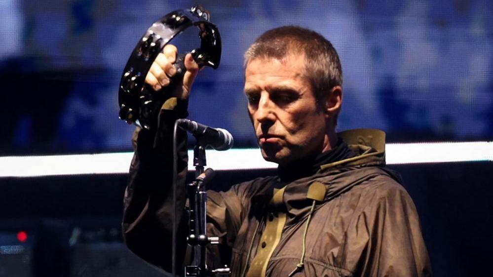 Liam Gallagher, wearing a raincoat, has a sombre expression as he holds up a tambourine during an Oasis reunion concert