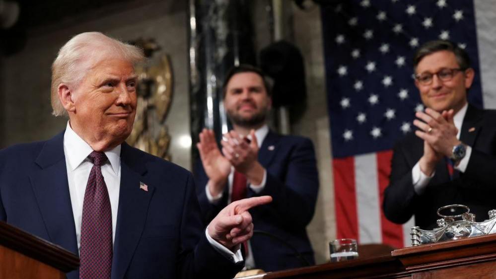 Donald Trump looks to his left, pointing with his index finger as JD Vance and Speaker of the House Mike Johnson stand behind him clapping their hands