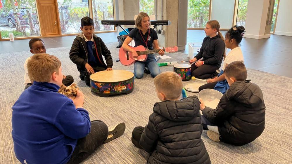 A group of children sit in a circle playing different instruments, including drums and tambourines, during a music therapy session. There is a woman in the centre of the circle leading the session. She is kneeling down and playing a guitar and singing.