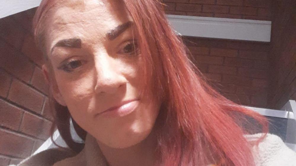 A selfie of a woman with red hair. She is sat outside in front of a red brick wall.