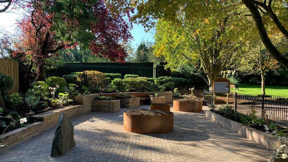 A paved garden with memorial rocks, large planters round the edge with new shrubs, large steel planters in the middle and benches up against the wall. Under large trees, blue sky and sunlight coming through the branches