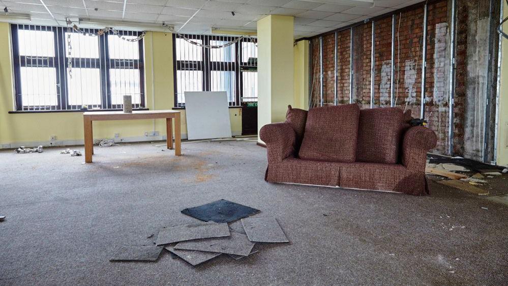 A rundown office unit in Rock House containing a disused sofa, table and carpet mats in the foreground. Leftover bunting can be seen hanging from the curtain rails. 