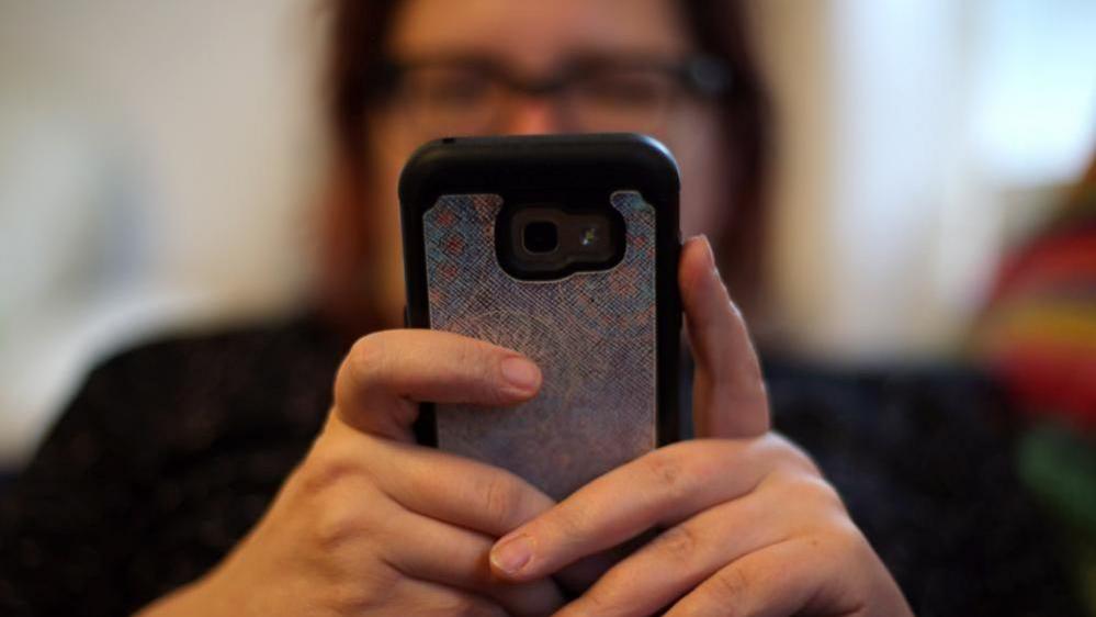 A woman wearing glasses, who is blurred, looks at a smartphone she is holding in her hands.