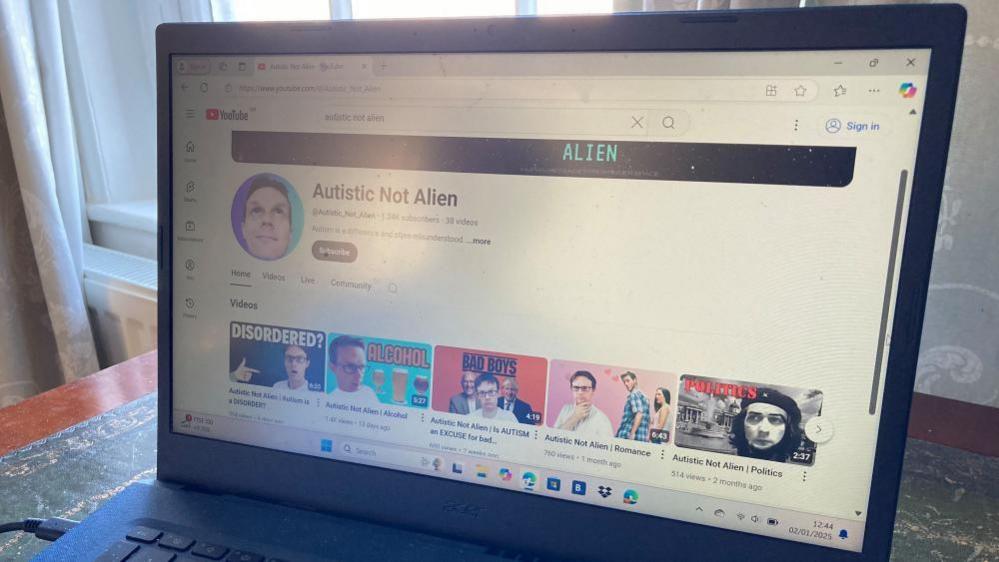 A computer screen that is on a desk in front of a window. The computer is open and on a YouTube page called "Autistic Not Alien". 