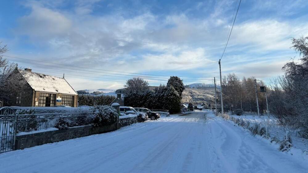 Snow covers a road with one house at the side of the road with two cars parked in the driveway. Snowy hills are in the distance.