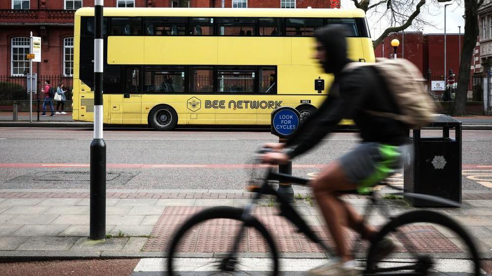 A yellow Bee Network double decker bus seen driving along a city centre road, with a cyclist in the foreground.