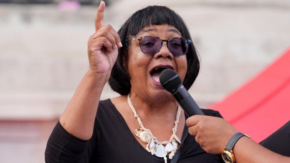 Labour MP Diane Abbott speaks into a microphone at a Trafalgar Square vigil staged to commemorate Jason Arday. She is dressed in black and wears dark glasses and white jewellery.