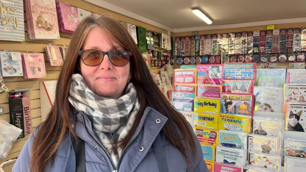 Mel Wright, has long brunette hair, is wearing tinted spectacles and a blue coat with black and white chequered scarf. She is standing inside her card and gift-wrap stall.