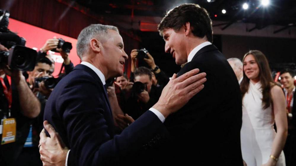 Mark Carney and Justin Trudeau embrace as a crowd of photographers take photos and Trudeau's daughter, who had introduced her father at the Liberal Party event, watch on. 