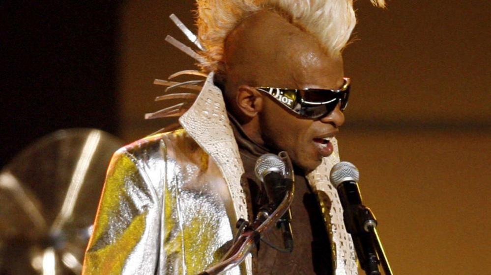  Funk music pioneer Sly Stone in a blond mohark and gold jacket, wearing sunglasses and singing into two microphones 