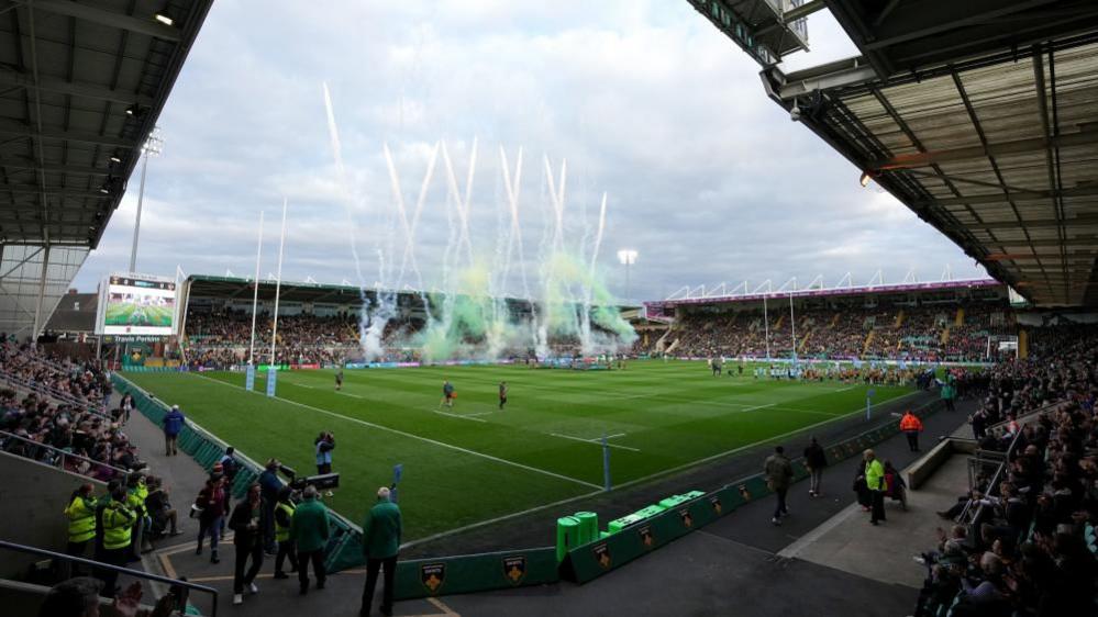 Spectators in stands on four sides of a rugby ground. Teams are walked out and smoke is billowing on one side of the pitch.