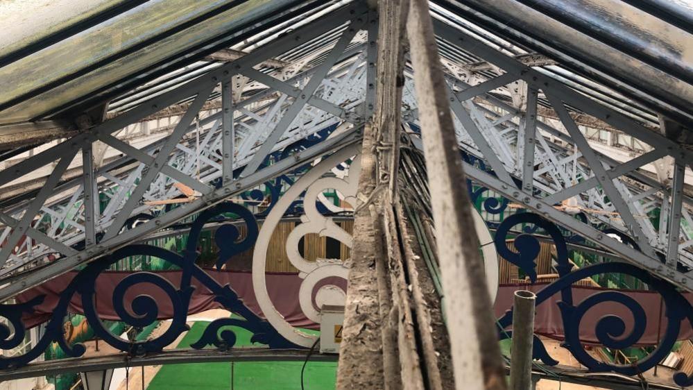 A view from the lantern, looking along the ridge of the main hall of the Great Yarmouth Winter Gardens, showing peeling paint and dust on some of the ironwork. Visible is the ornate structure, and trusses, with the green carpet and a brown curtain hanging down in the mid distance.