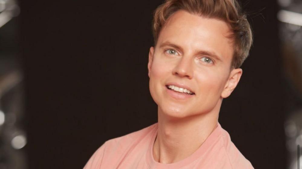 A professional headshot of a young man with blond hair and green eyes