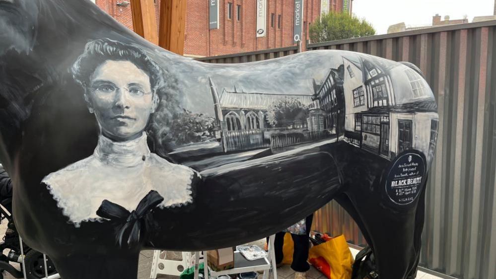 Black Beauty author Anna Sewell, painted on the side of a black fibre glass horse statue, with other images depicting the house where she was born, a timber-fronted property, which is adjacent to Great Yarmouth Minster - a large parish church, with copper roof and arched gothic windows. The picture is taken inside Great Yarmouth Market, with fencing and Market Gates Shopping Centre behind.