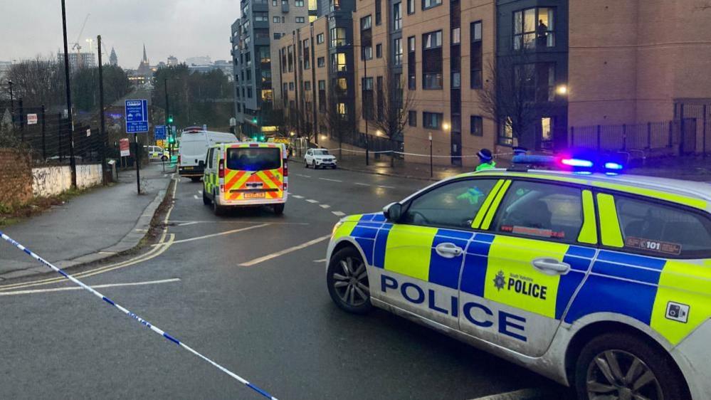 A police car and police van on a road next to multiple apartment buildings. The road has been cordoned off with police tape.