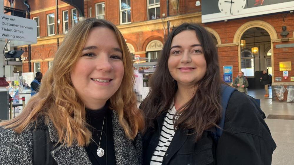 Rebecca Sheridan, left,  with auburn hair, wearing a grey mottled coat and black jersey wearing a silver necklace pendant, and Leia Henderson, has black hair, dressed in a black and white striped top, with black jacket and blue rucksacks trap over her shoulder. They are standing on the concourse at Norwich station near the station clock.