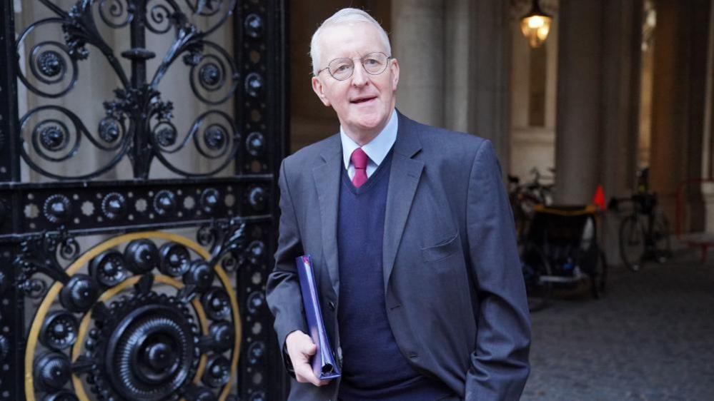 Benn is looking at the camera while walking out of a building. He is wearing a dark grey suit with a navy jumper underneath. He has a red tie on and light coloured shirt. He is holding a blue file under his arm.