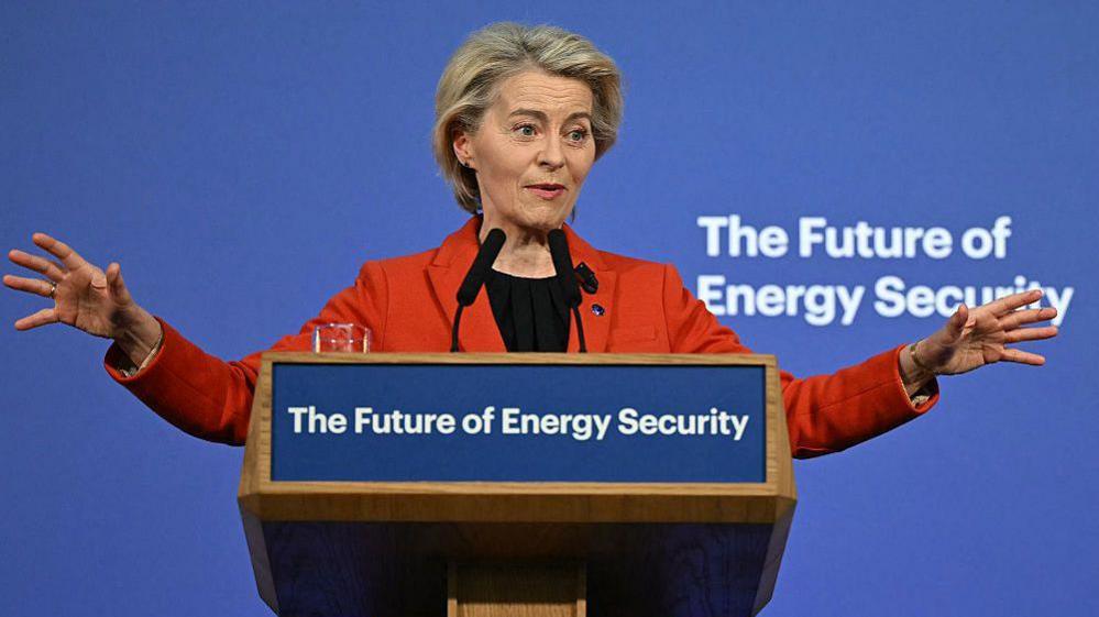 Një grua me një xhaketë portokalli flet në një podium