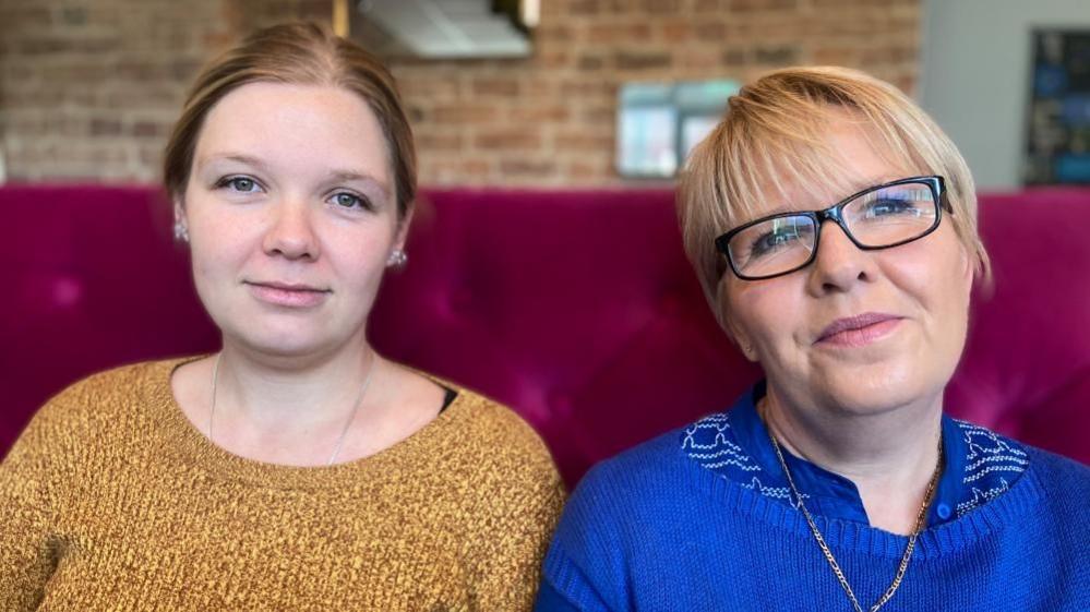 Samantha and Maddie Nunn sit side by side on a pink sofa. Maddie has sandy blonde hair pulled back in a centre part and wears a gold jumper. Samantha has blonde hair in a pixie crop cut and wears glasses and a blue jumper. 