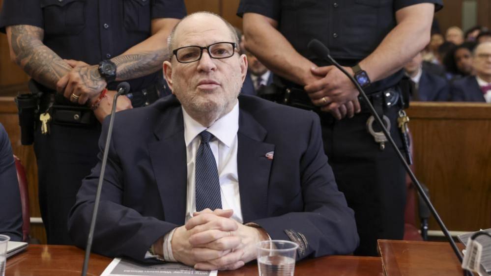 Harvey Weinstein is wearing a blue suit and tie and sitting in a courtroom. he has his hands clasped and is wearing a US flag pin at his lapel. Two police officers stand behind him.