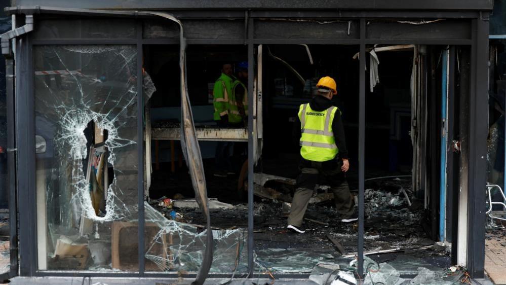 A man wearing a high vis vest and yellow hard hat walks through the charred remains of a building