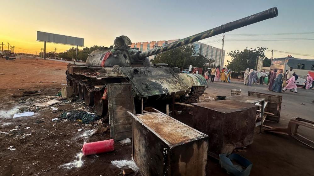 A military tank is standing on the side of a road in Wad Mdani, with people in civilian clothes nearby