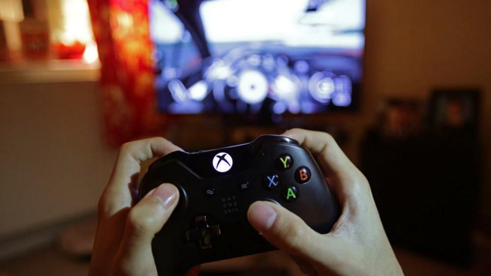 Two hands holding a black video game controller. The letters X, Y, B, A, are visible on the gadget. In the background is a blurred screen.