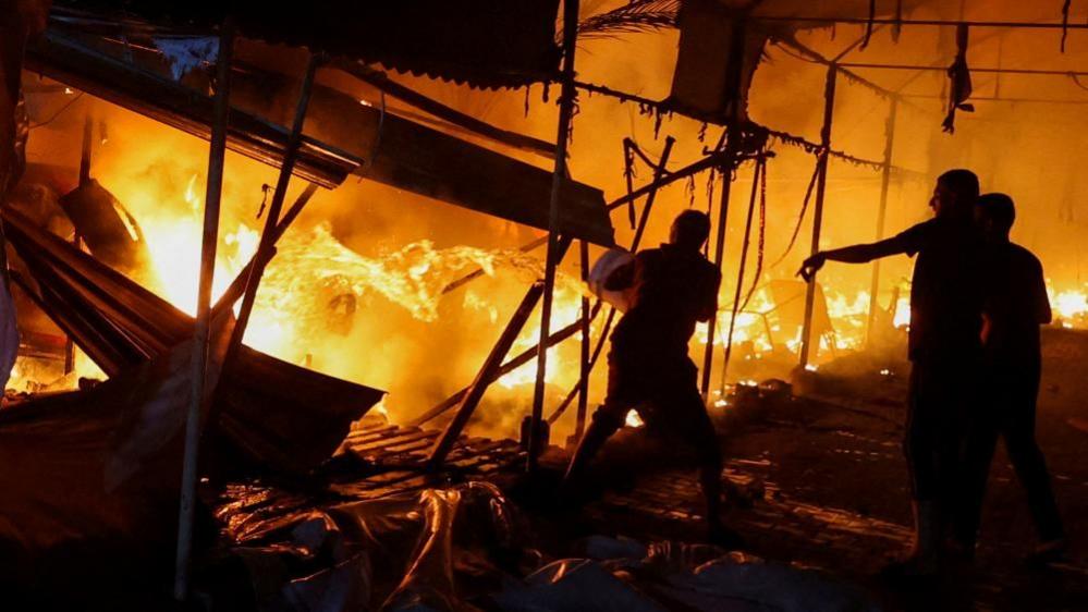 Three Palestinian men throw water from a bucket, as they try to put out a wall of flames at the al-Aqsa complex after an Israeli air strike. What looks like corrugated iron and debris can be seen collapsed in the background.