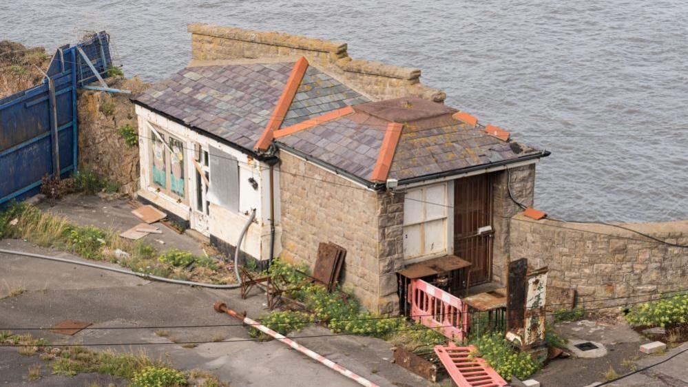 The derelict Toll House at Birnbeck Pier, Weston-super-Mare. The windows are boarded and surrounding area overgrown with weeds. 