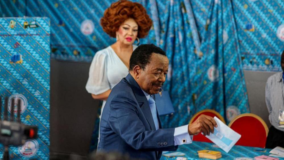 Biya putting his ballot into the box, with his wife standing behind him. There is a bright blue background.