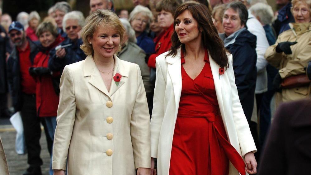 Kathryn Apanowicz smiles while walking past crowds of people. She is wearing a cream-coloured coat with a poppy pinned to it. She is walking alongside Carol Vorderman, who is wearing a red dress and cream-coloured coat as well as a poppy.