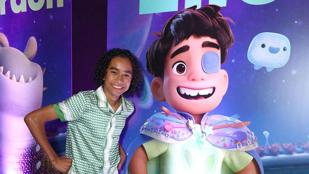 Actor Yonas Kibreab posing next to a poster of Elio, featuring the titular character that he plays.