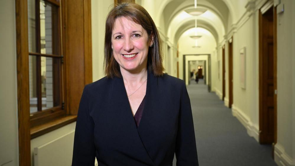 A woman in a black jacket with brown hair smiles in a corridor in front of an internal window.